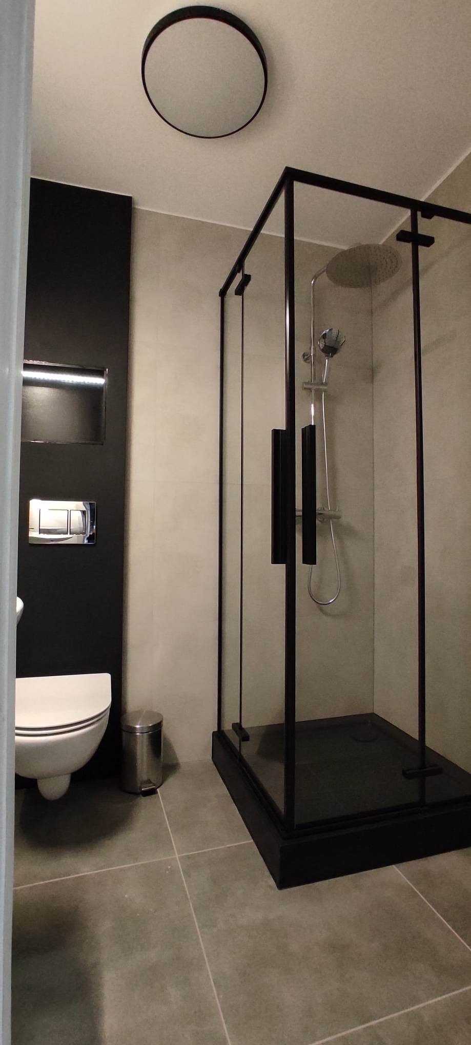 Nowoczesna łazienka z kabiną prysznicową typu walk-in z czarnymi profilami, wiszącą toaletą i okrągłym lustrem w suficie.