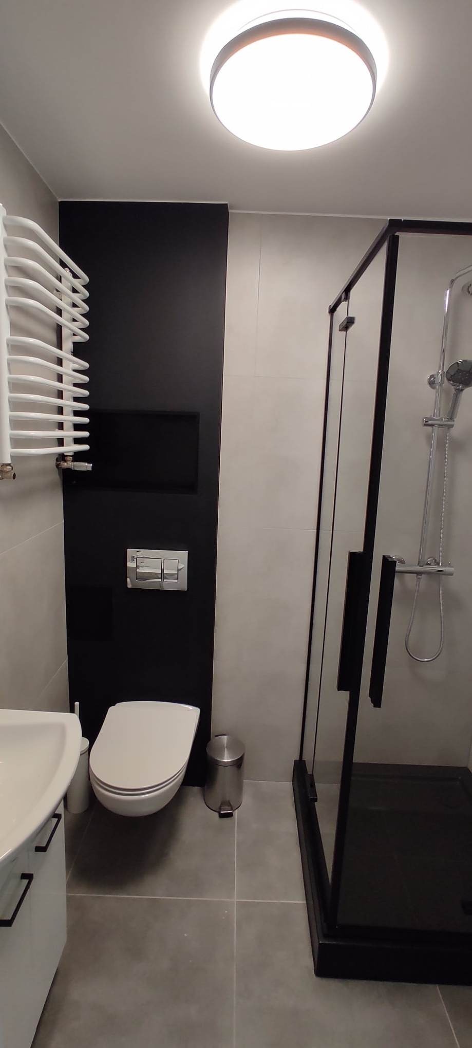 Nowoczesna łazienka z czarną ścianą akcentującą, kabiną prysznicową z czarną ramą, podwieszaną toaletą i grzejnikiem drabinkowym.