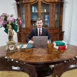 Młody mężczyzna w kraciastej marynarce siedzi za dużym, okrągłym biurkiem z laptopem Dell, obok niego statuetka Temidy, zegar, akta i kodeksy, a w tle ozdobny kredens.