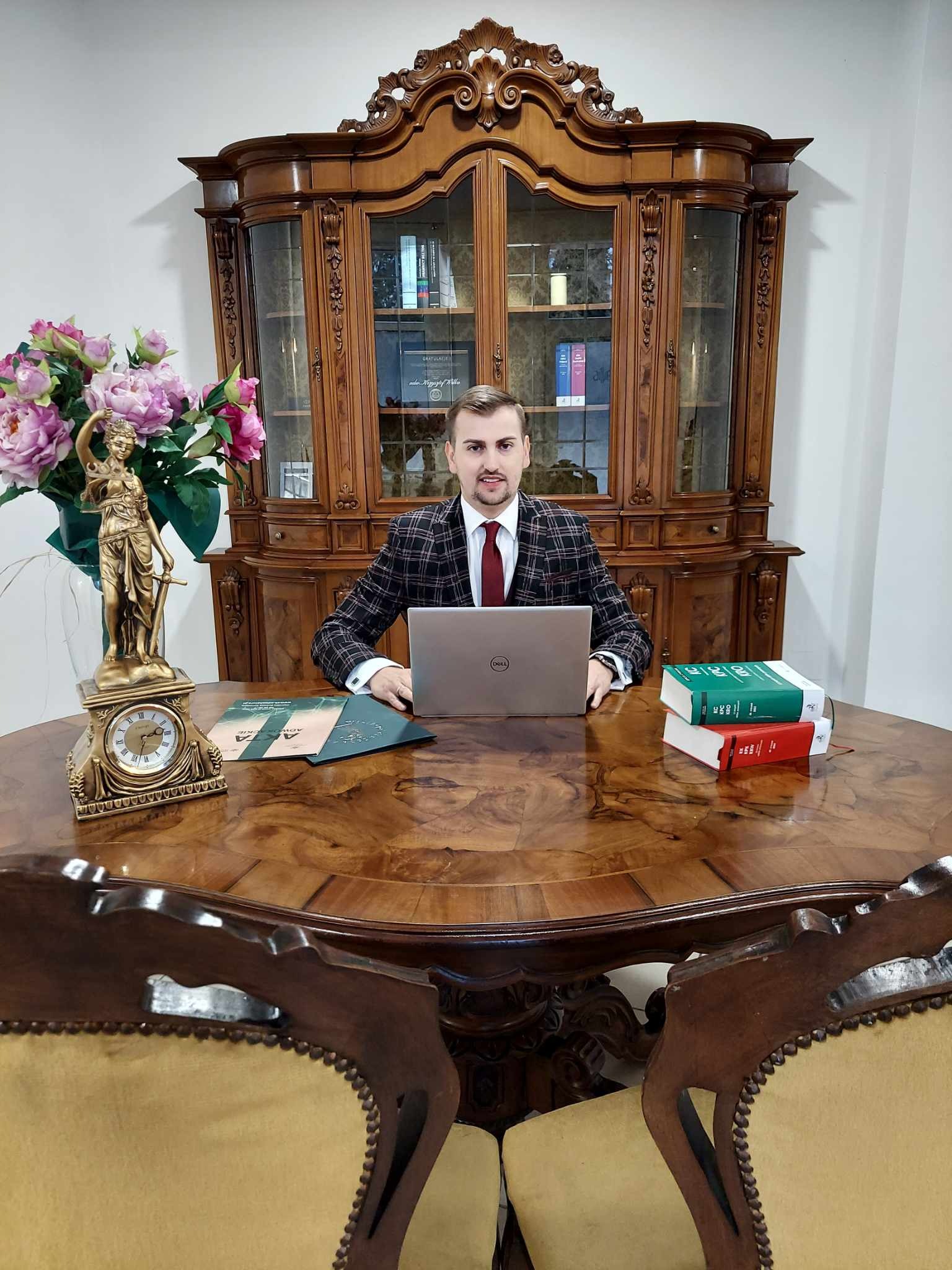 Młody mężczyzna w kraciastej marynarce siedzi za dużym, okrągłym biurkiem z laptopem Dell, obok niego statuetka Temidy, zegar, akta i kodeksy, a w tle ozdobny kredens.