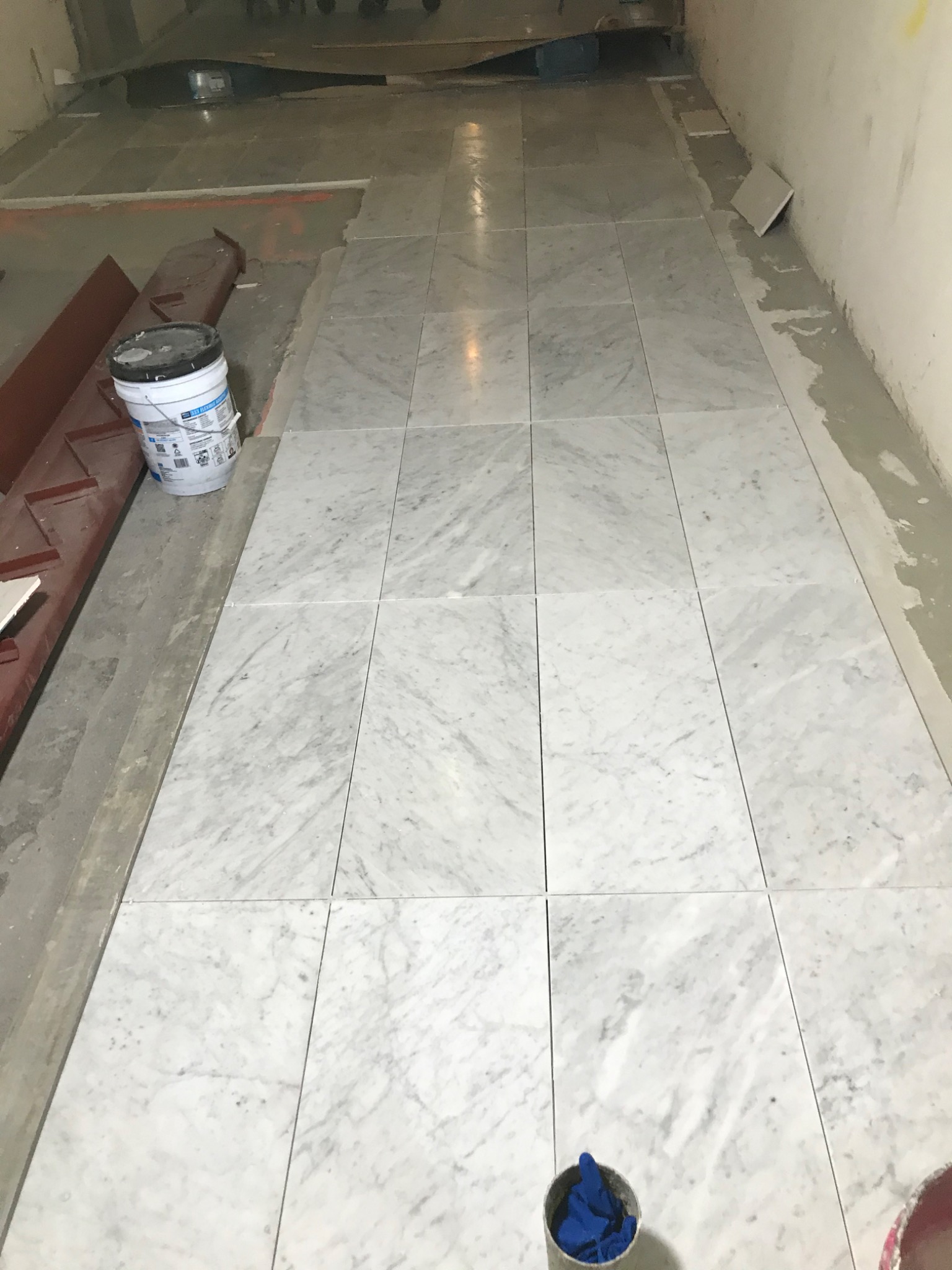 Ułożone, białe marmurowe płytki podłogowe podczas remontu, widoczny wiadro z klejem i narzędzia.