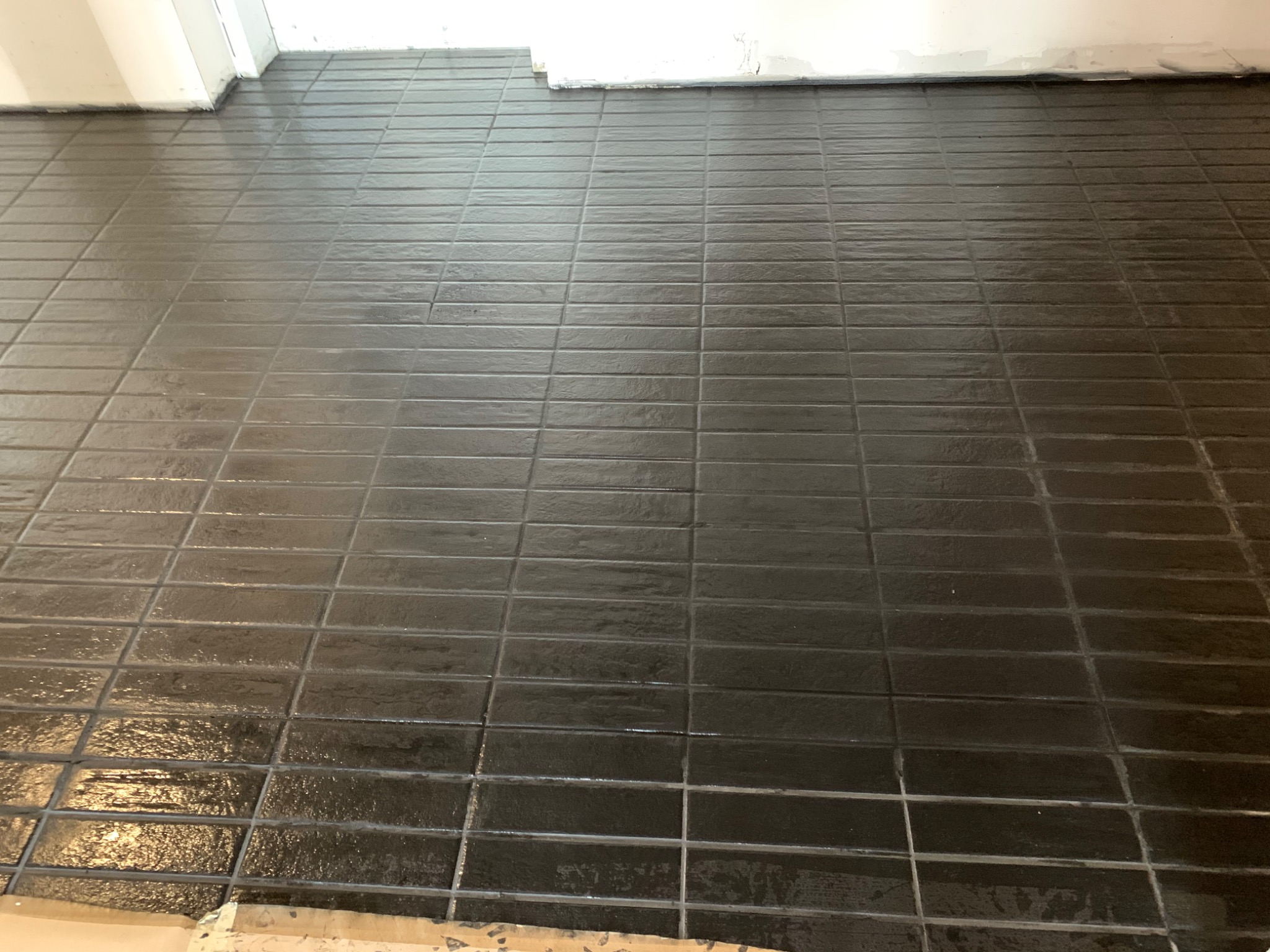 Błyszcząca, ułożona w cegiełkę, ciemnoszara podłoga z płytek ceramicznych, widok z góry, przy białej ścianie.