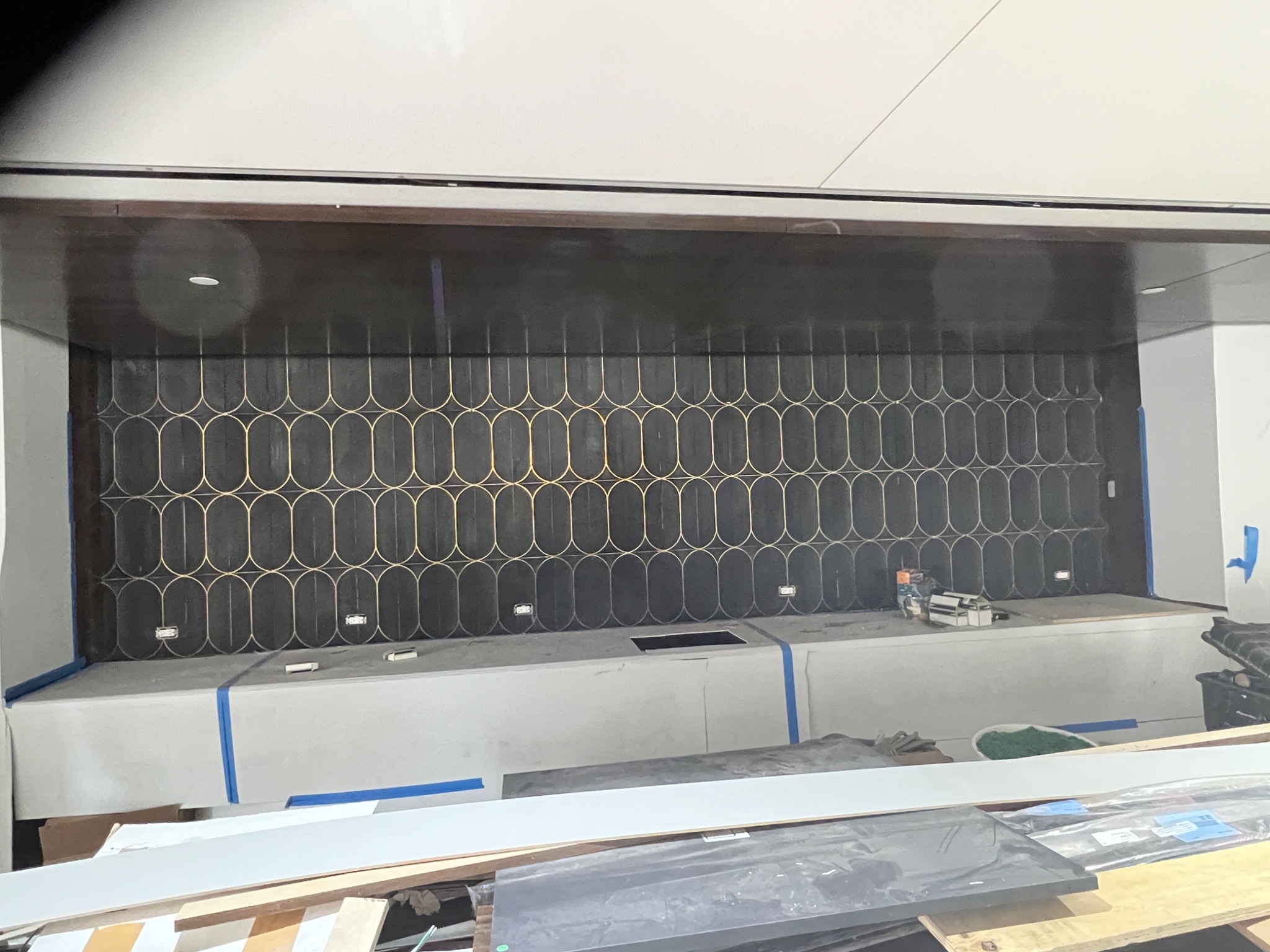 Ściana z ciemnych płytek z geometrycznym, złotym wzorem w trakcie montażu, zabezpieczona niebieską taśmą malarską, widoczny blat roboczy i narzędzia.