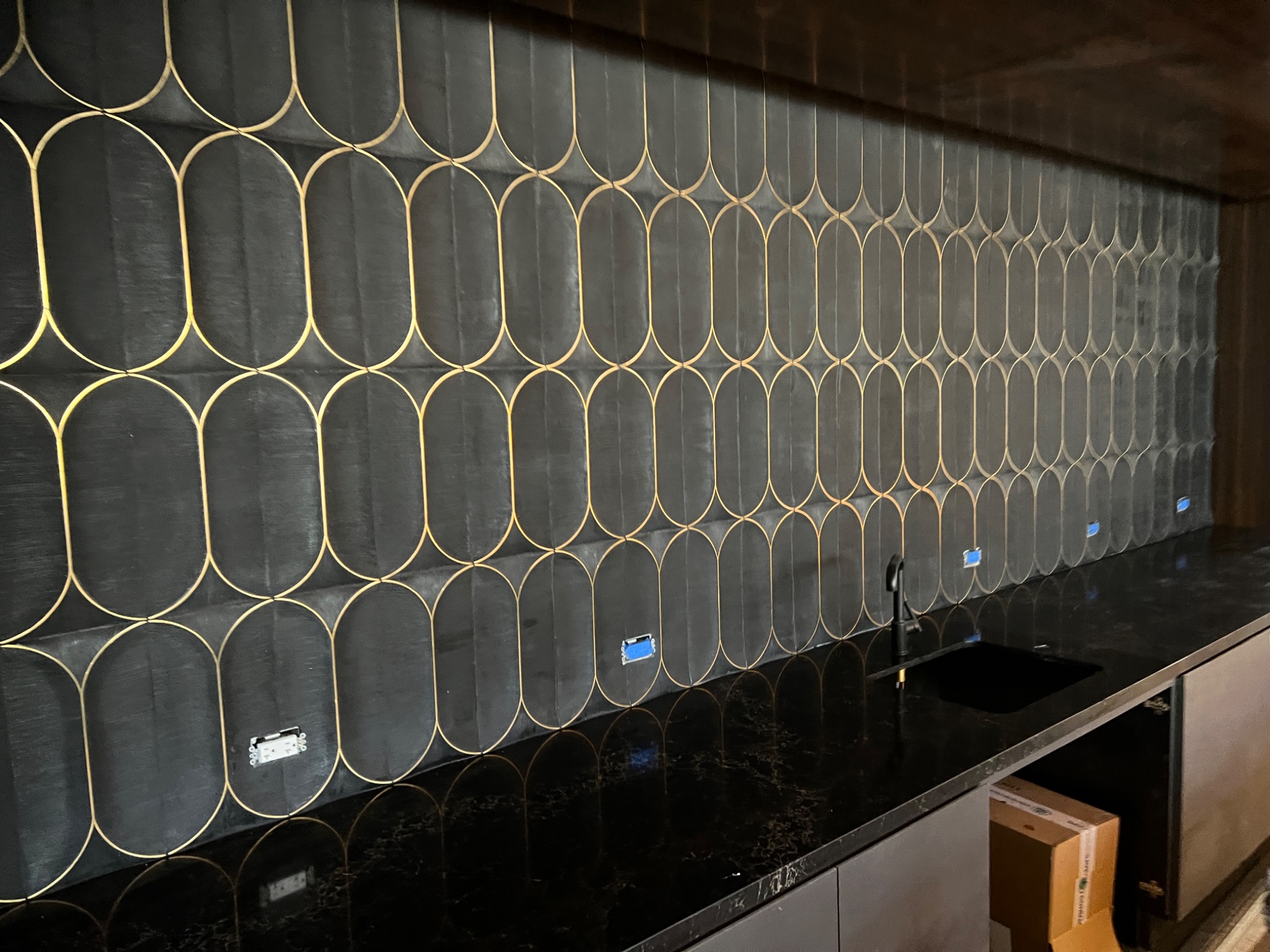 Czarna ściana z owalnych płytek z złotymi obwódkami nad czarnym blatem z umywalką, widoczne gniazdka elektryczne.