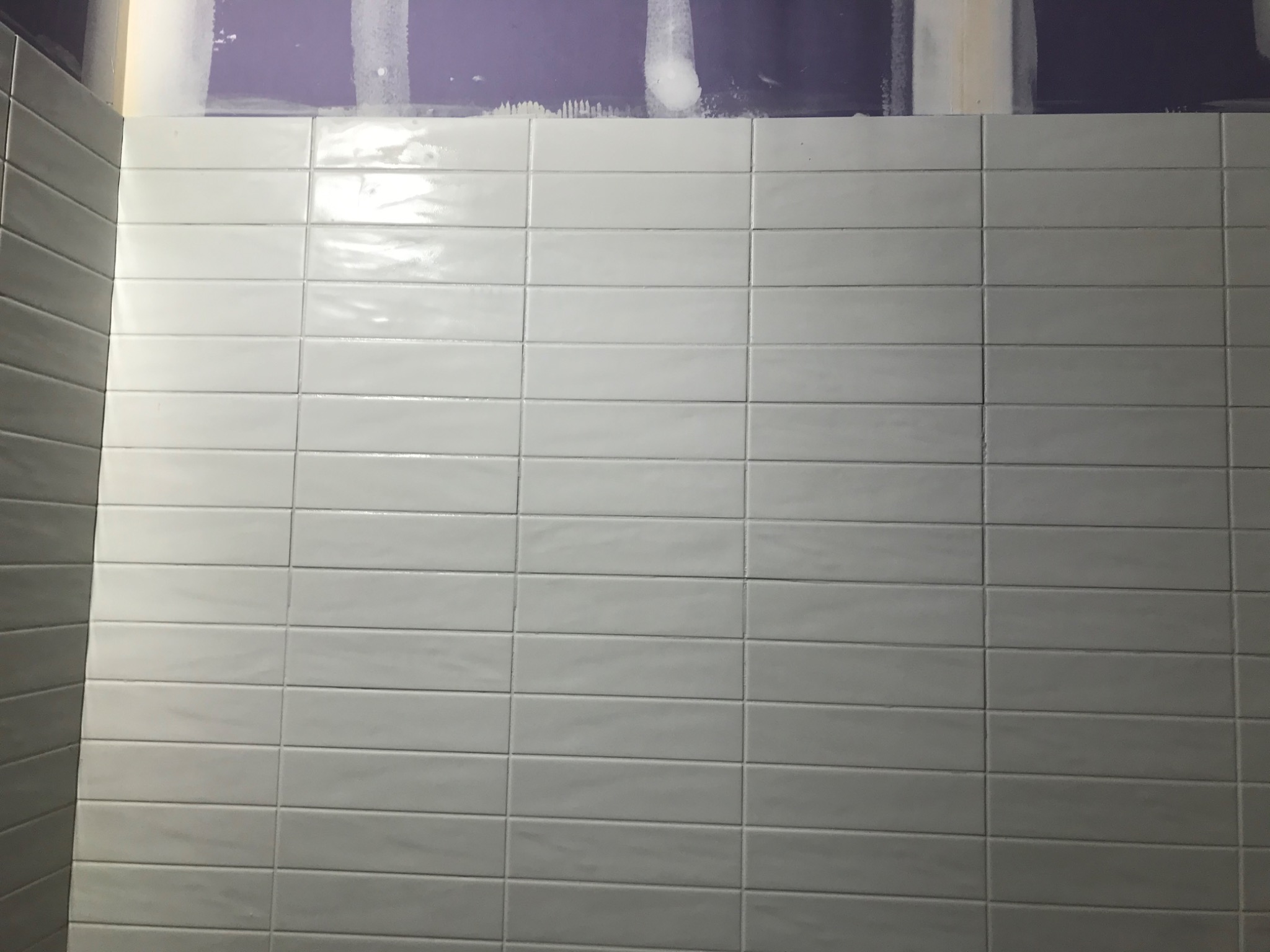 Ściana łazienki wyłożona białymi, prostokątnymi płytkami, z widocznymi śladami kleju na górze i po bokach, sugerującymi świeżo zakończony montaż.