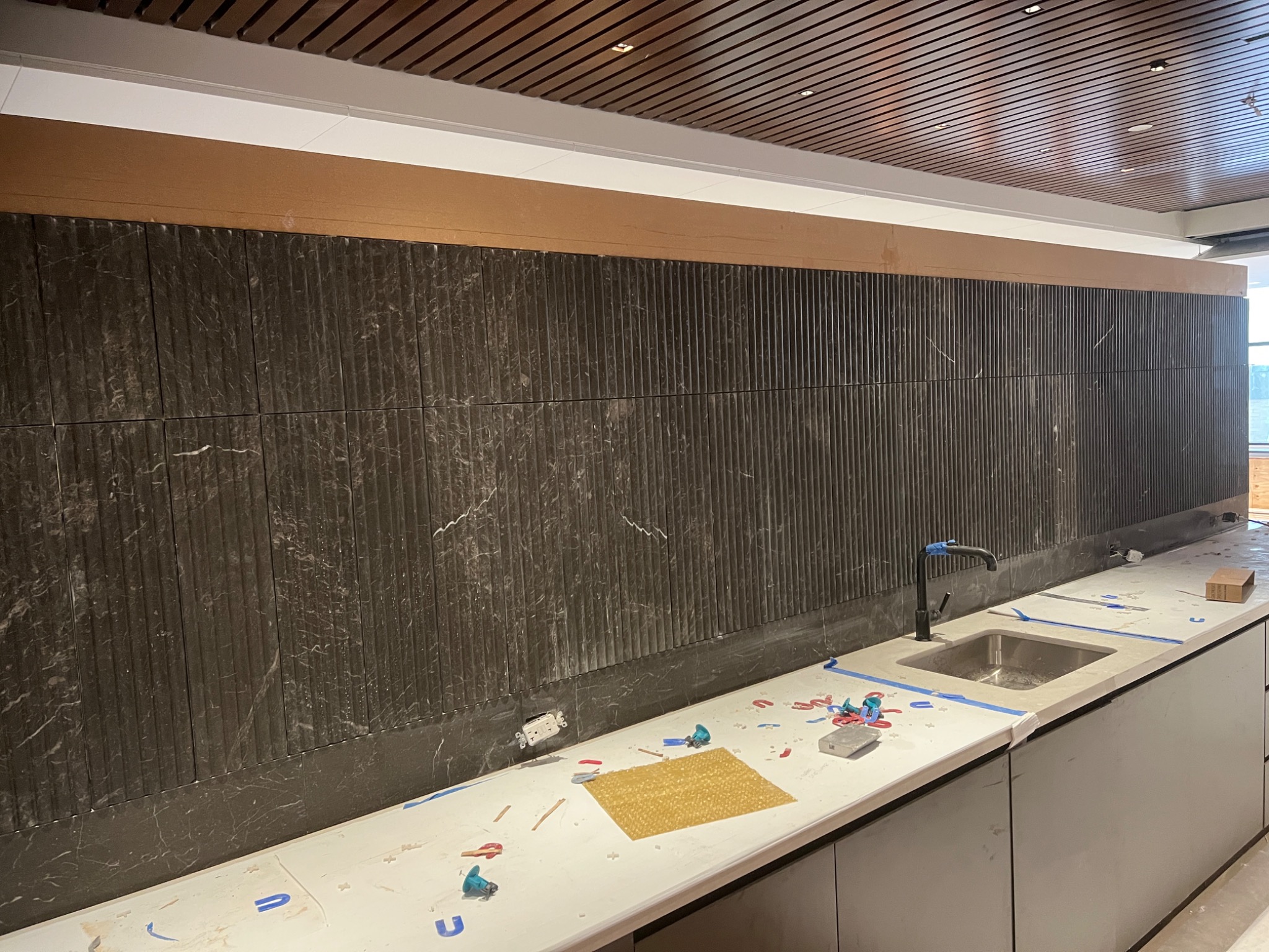 Montaż płytek z efektem marmuru na ścianie w kuchni, widoczny blat z umywalką i akcesoria glazurnicze, sufit z drewnianymi panelami.