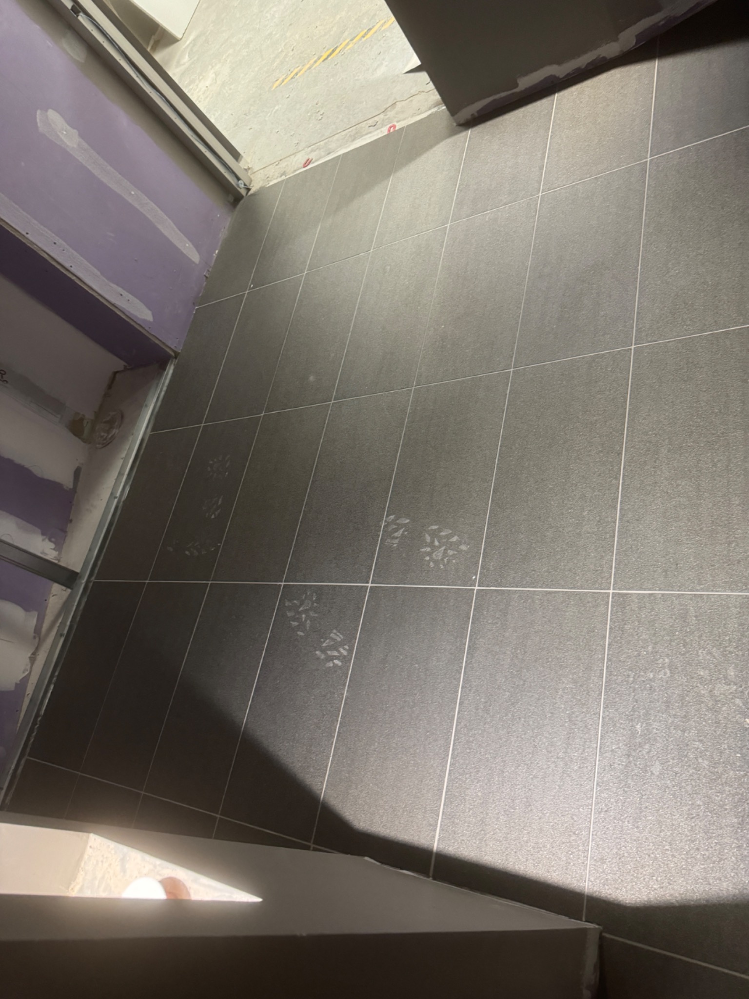 Ułożone szare płytki podłogowe z widocznymi śladami butów, przy ścianie z płyt gipsowo-kartonowych w trakcie montażu.