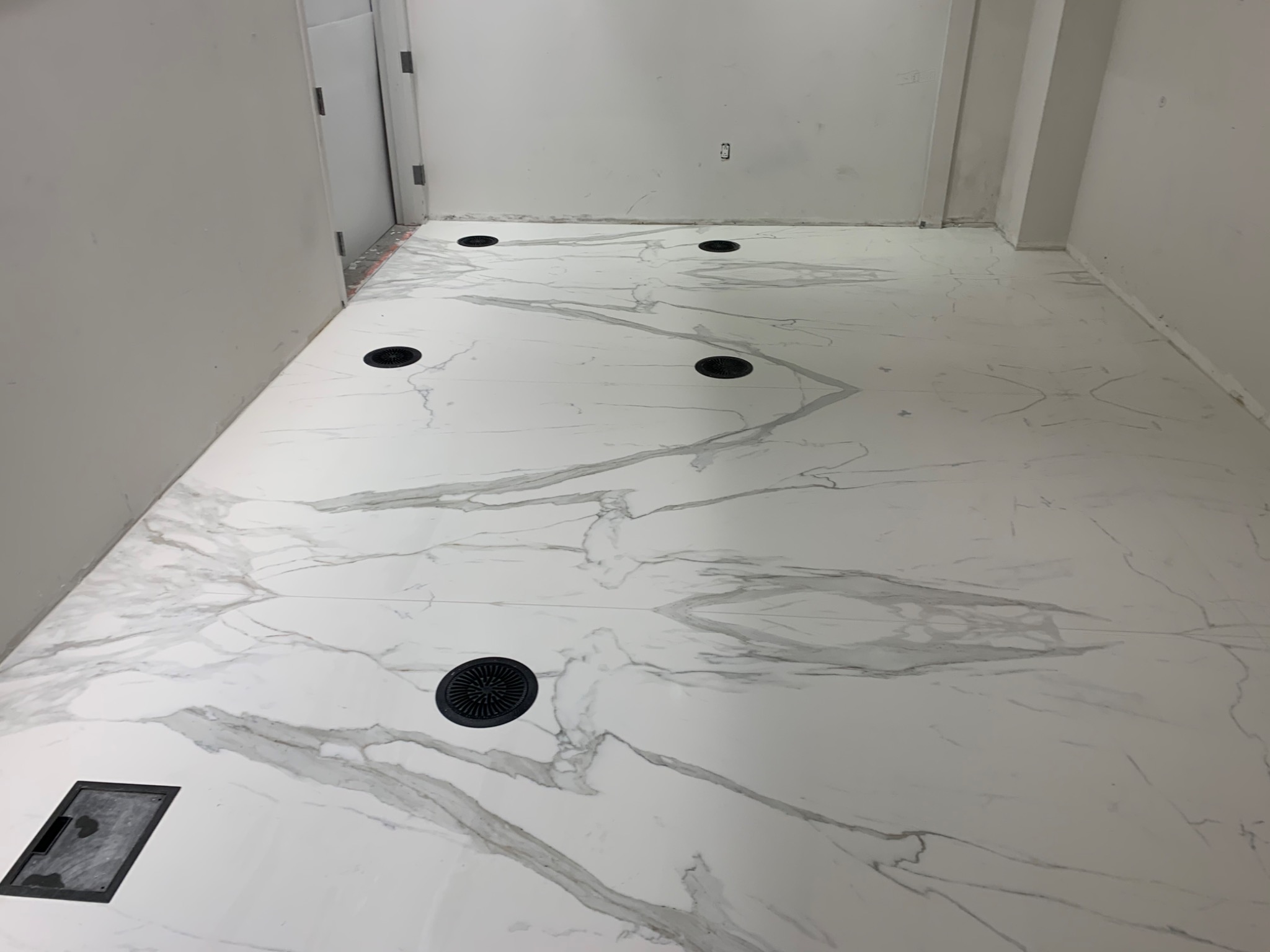 Wykończona podłoga z białych płytek imitujących marmur z czarnymi odpływami liniowymi i punktowymi w nowym pomieszczeniu.