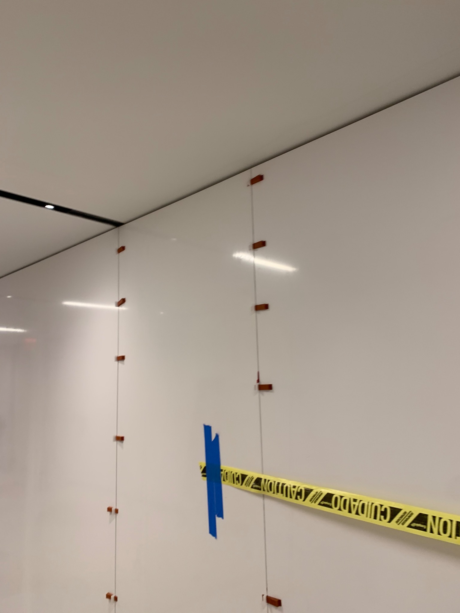 Białe, błyszczące płytki ścienne w trakcie montażu z użyciem brązowych krzyżyków dystansowych i taśmy ostrzegawczej z napisem 'Caution/Cuidado' przytrzymywanej niebieską taśmą.