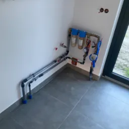 Szare rury doprowadzające wodę do filtrów i wodomierzy zamontowanych na ścianie w rogu pomieszczenia z szarą podłogą.