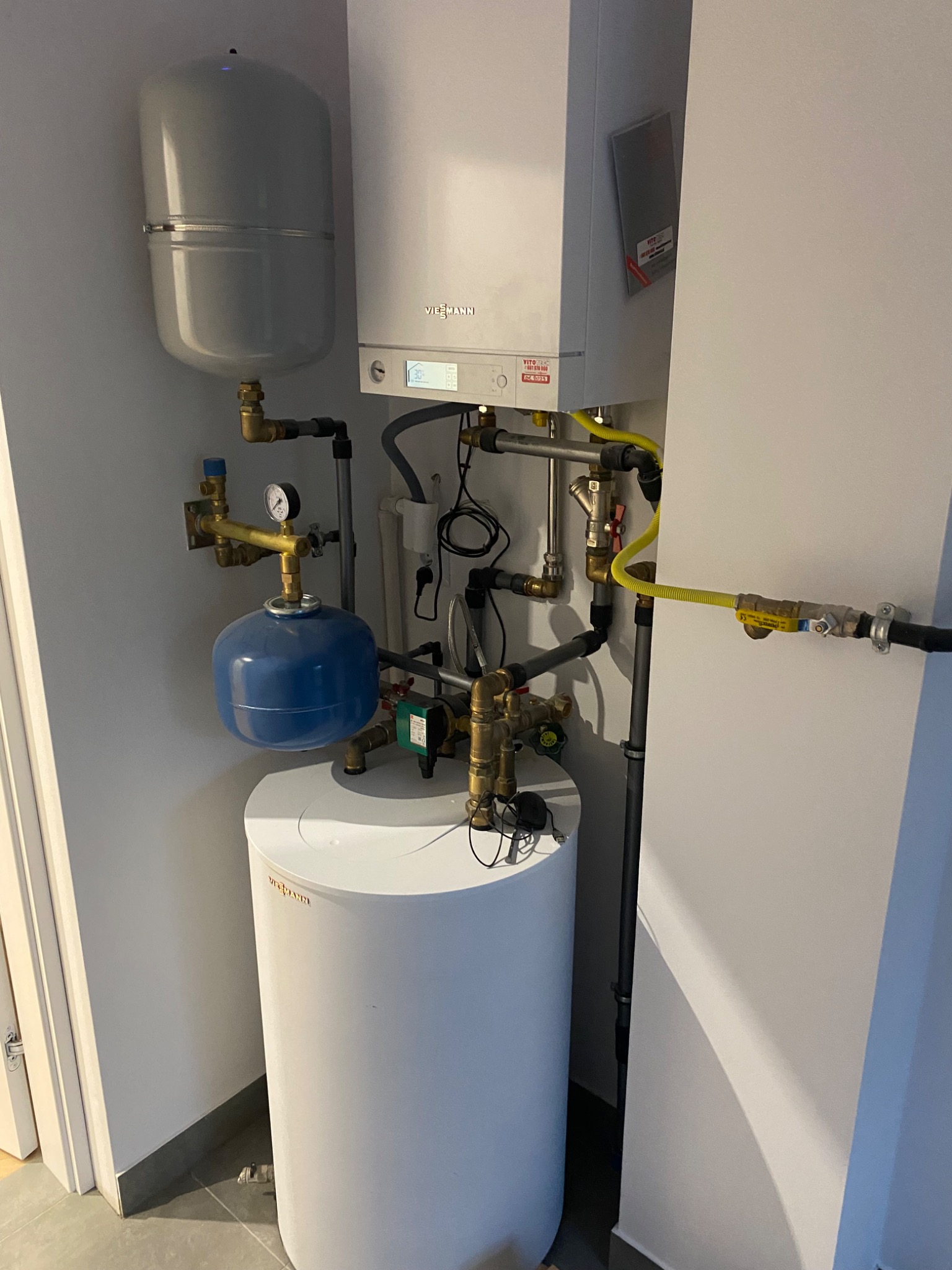 Nowoczesny, biały piec gazowy Viessmann z podłączonymi rurami miedzianymi i żółtą rurą gazową, widoczny zbiornik wyrównawczy i zasobnik ciepłej wody, w minimalistycznym wnętrzu.