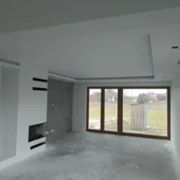 Surowe wnętrze w trakcie prac wykończeniowych: białe ściany, sufit podwieszany z oświetleniem punktowym, dekoracyjna ściana z szarego kamienia z czarnymi liniami, kominek z czarną obudową, duże...