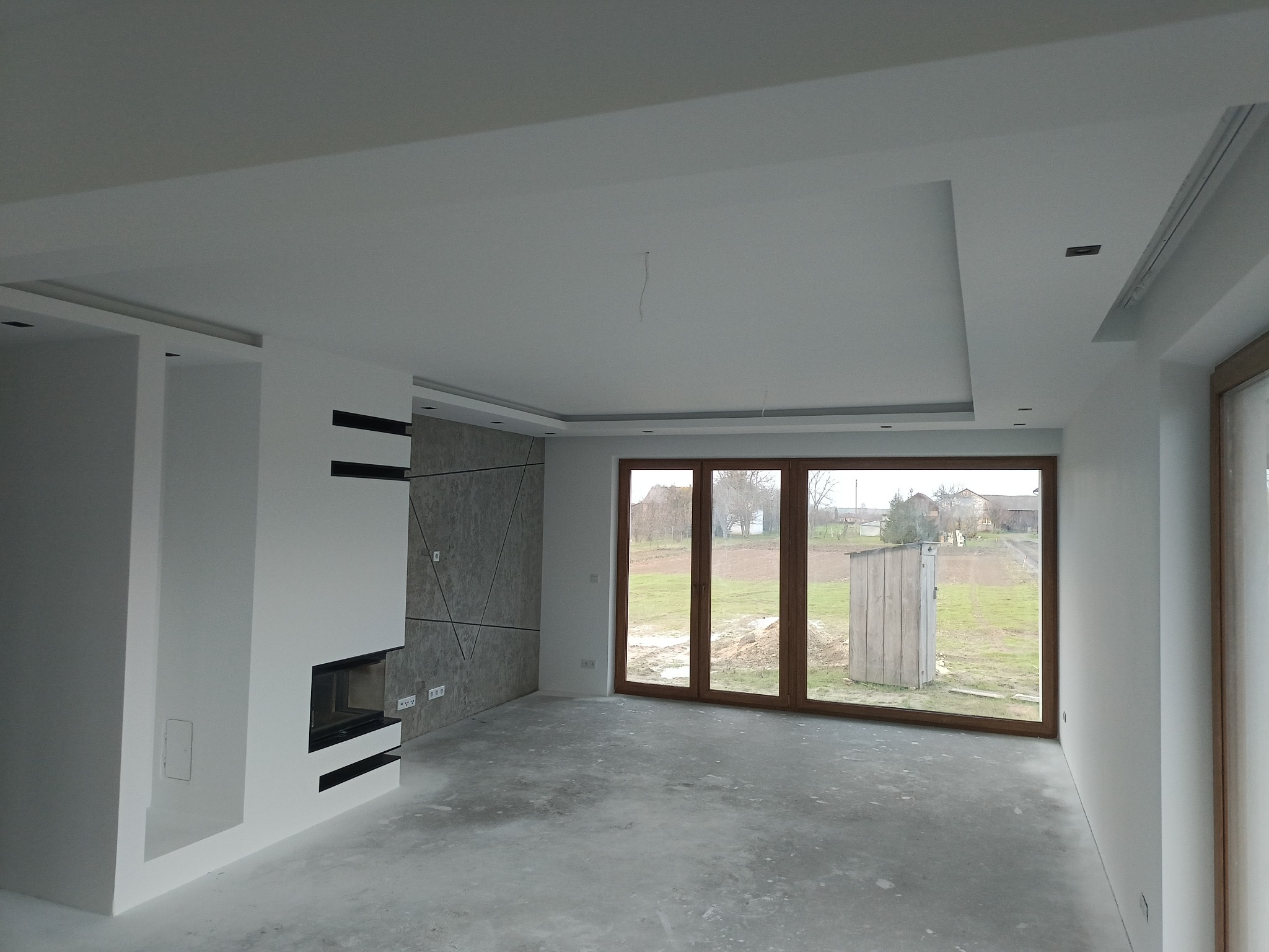 Surowe wnętrze w trakcie prac wykończeniowych: białe ściany, sufit podwieszany z oświetleniem punktowym, dekoracyjna ściana z szarego kamienia z czarnymi liniami, kominek z czarną obudową, duże...