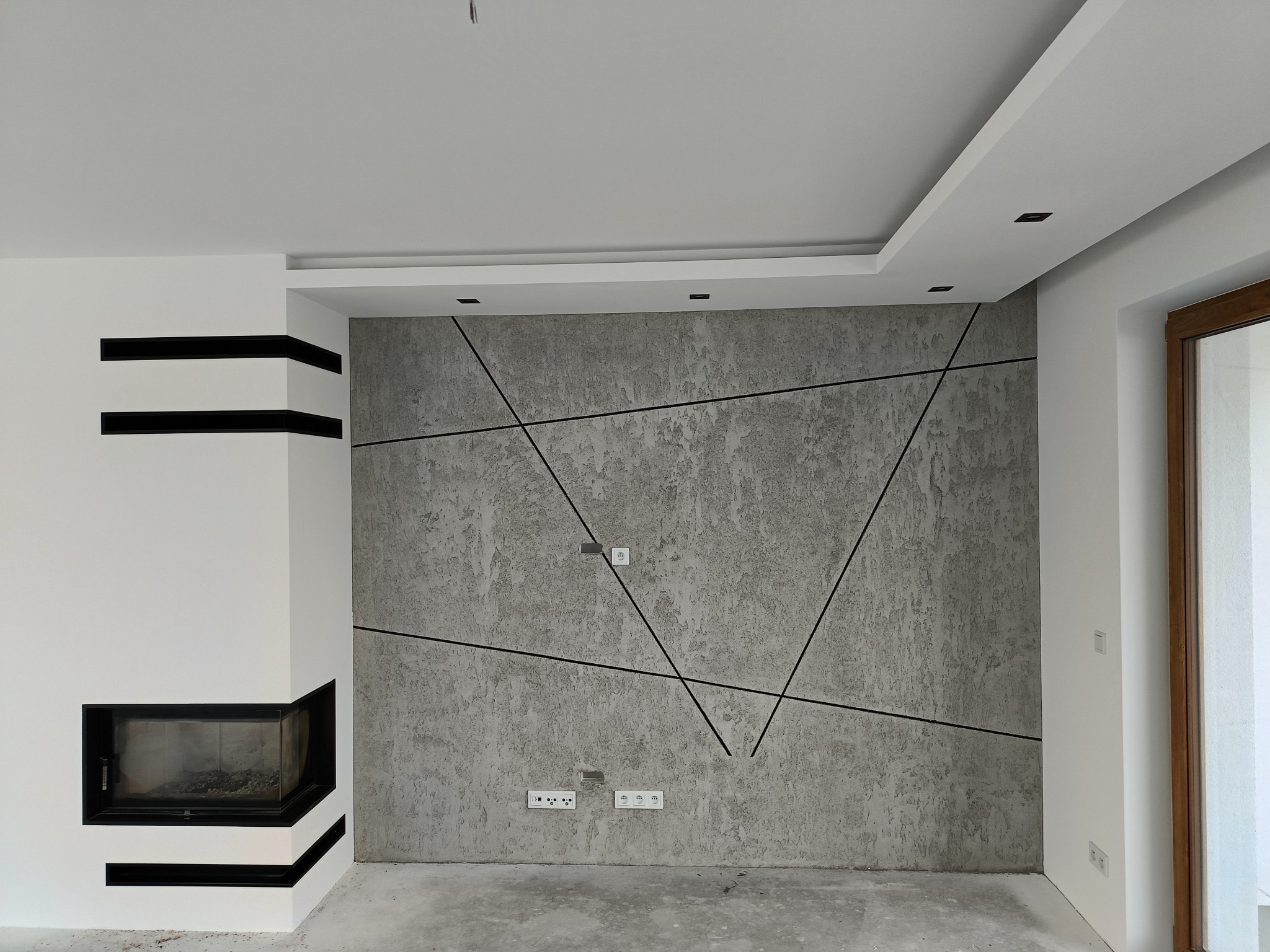 Szara ściana dekoracyjna z geometrycznym wzorem z czarnych linii, gładka biała ściana z minimalistycznym kominkiem z czarnymi akcentami, sufit z oświetleniem punktowym, wnętrze w trakcie wykańczania.