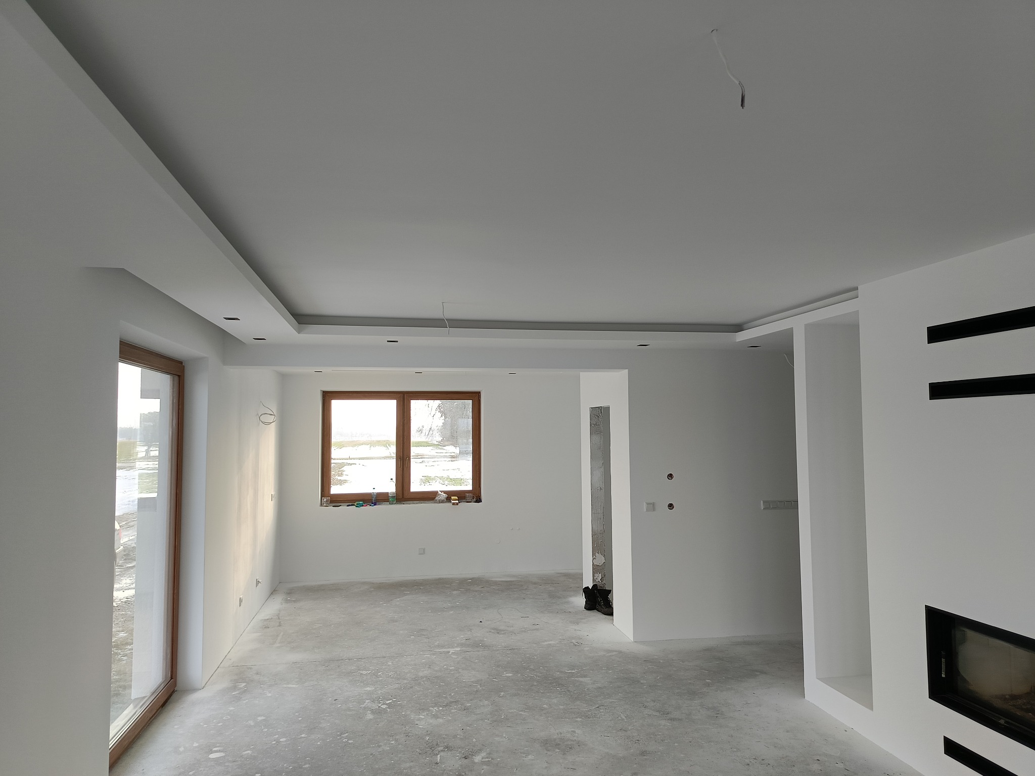 Wnętrze w trakcie wykańczania, widoczne gładkie, białe ściany, sufit z podwieszanym elementem i wnęką na oświetlenie, surowa podłoga, okna z drewnianą ramą oraz fragment kominka z czarną obudową.