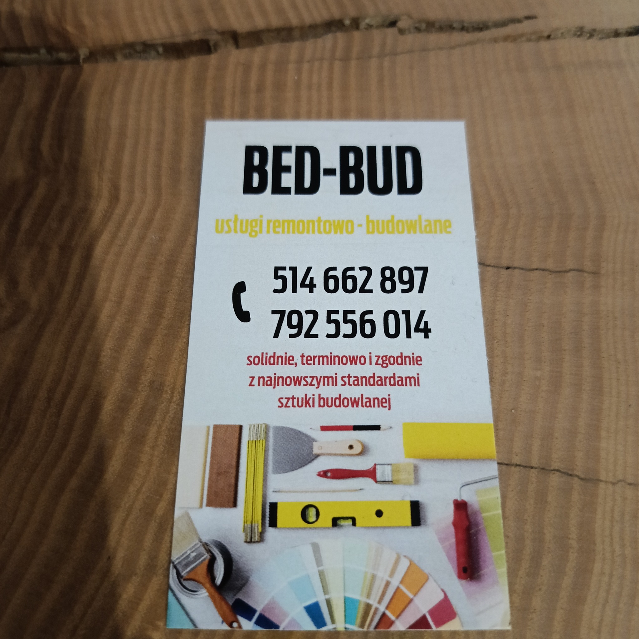 Wizytówka firmy remontowo-budowlanej BED-BUD z numerami telefonów, leżąca na drewnianej powierzchni. Na wizytówce grafika przedstawiająca narzędzia budowlane, takie jak pędzel, szpachelka...