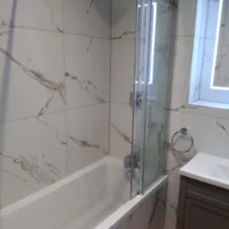 Nowoczesna łazienka z wanną i przeszklonym parawanem nawannowym, wykończona białymi płytkami imitującymi marmur z szarymi żyłami, podświetlane lustro nad umywalką.