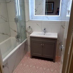 GREENKRIS - Nowoczesna łazienka po remoncie: białe marmurowe płytki ścienne, różowa podłoga z geometrycznym wzorem, szafka pod umywalką w kolorze taupe, oświetlone lustro, kabina prysznicowa ze szklanym panelem.