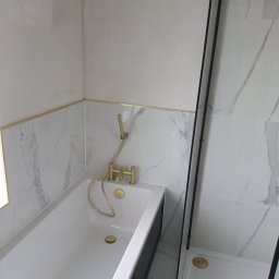 GREENKRIS - Nowoczesna łazienka z białą wanną, złotą armaturą i kabiną prysznicową z czarnymi profilami, wykończona marmurowymi płytkami.