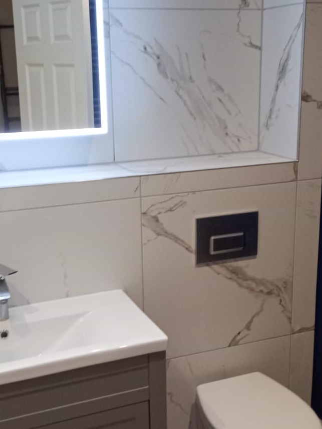 Wykończona łazienka z umywalką na szarej szafce, podświetlanym lustrem i płytkami imitującymi marmur z czarnym przyciskiem spłuczki.