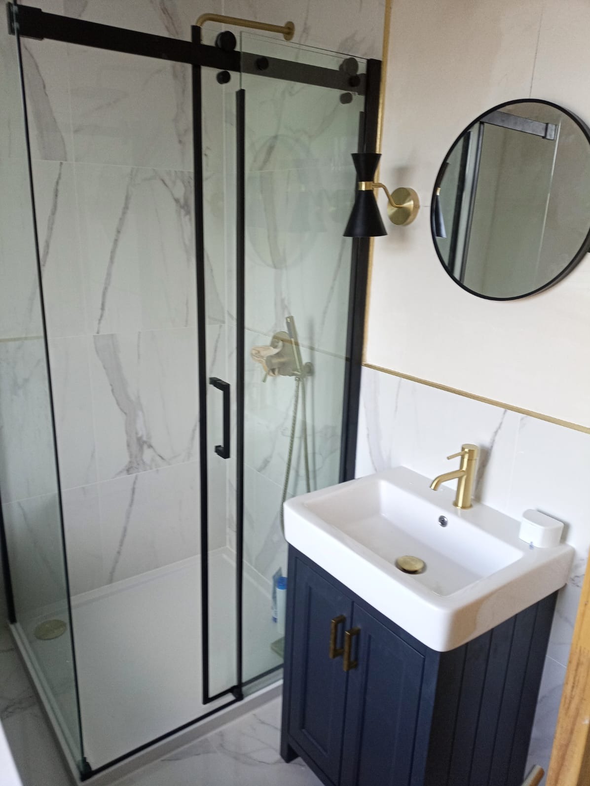Nowoczesna łazienka z kabiną prysznicową w czarnej ramie, umywalką na szafce w kolorze granatowym i złotą armaturą, oświetlona czarno-złotym kinkietem.