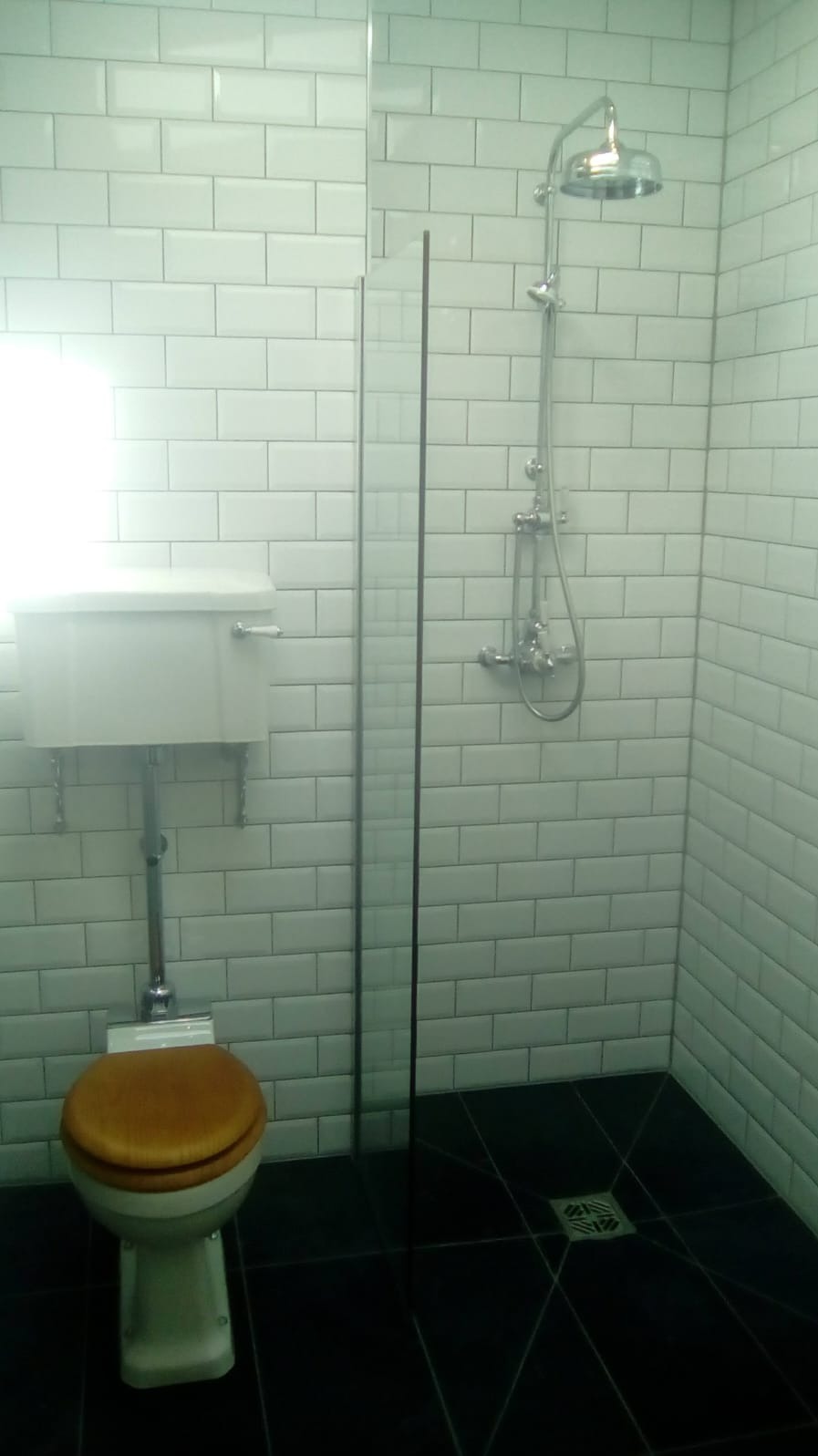 Wykończona łazienka z białymi, prostokątnymi płytkami na ścianach, czarną podłogą, szklaną kabiną prysznicową i toaletą ze spłuczką oraz drewnianą deską sedesową.