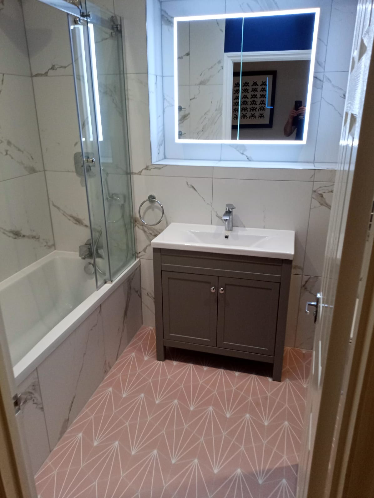 Nowoczesna łazienka po remoncie: białe marmurowe płytki ścienne, różowa podłoga z geometrycznym wzorem, szafka pod umywalką w kolorze taupe, oświetlone lustro, kabina prysznicowa ze szklanym panelem.