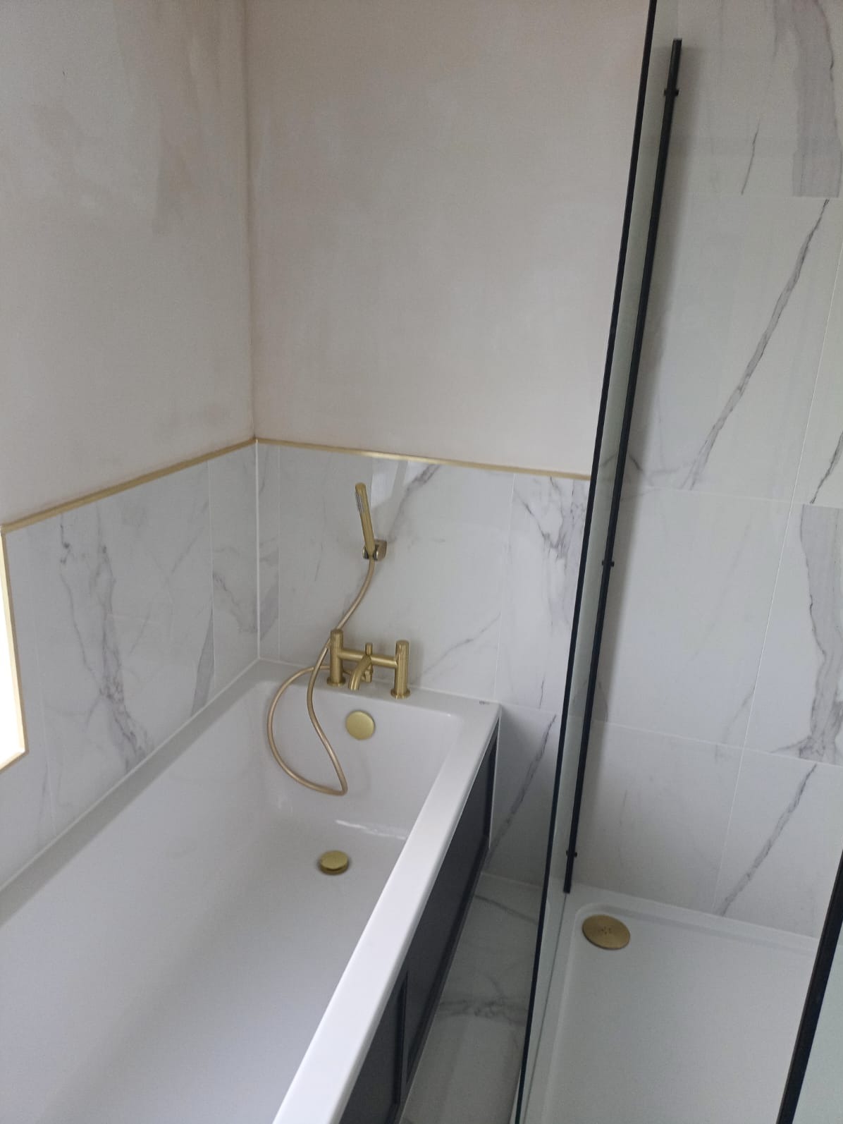 Nowoczesna łazienka z białą wanną, złotą armaturą i kabiną prysznicową z czarnymi profilami, wykończona marmurowymi płytkami.