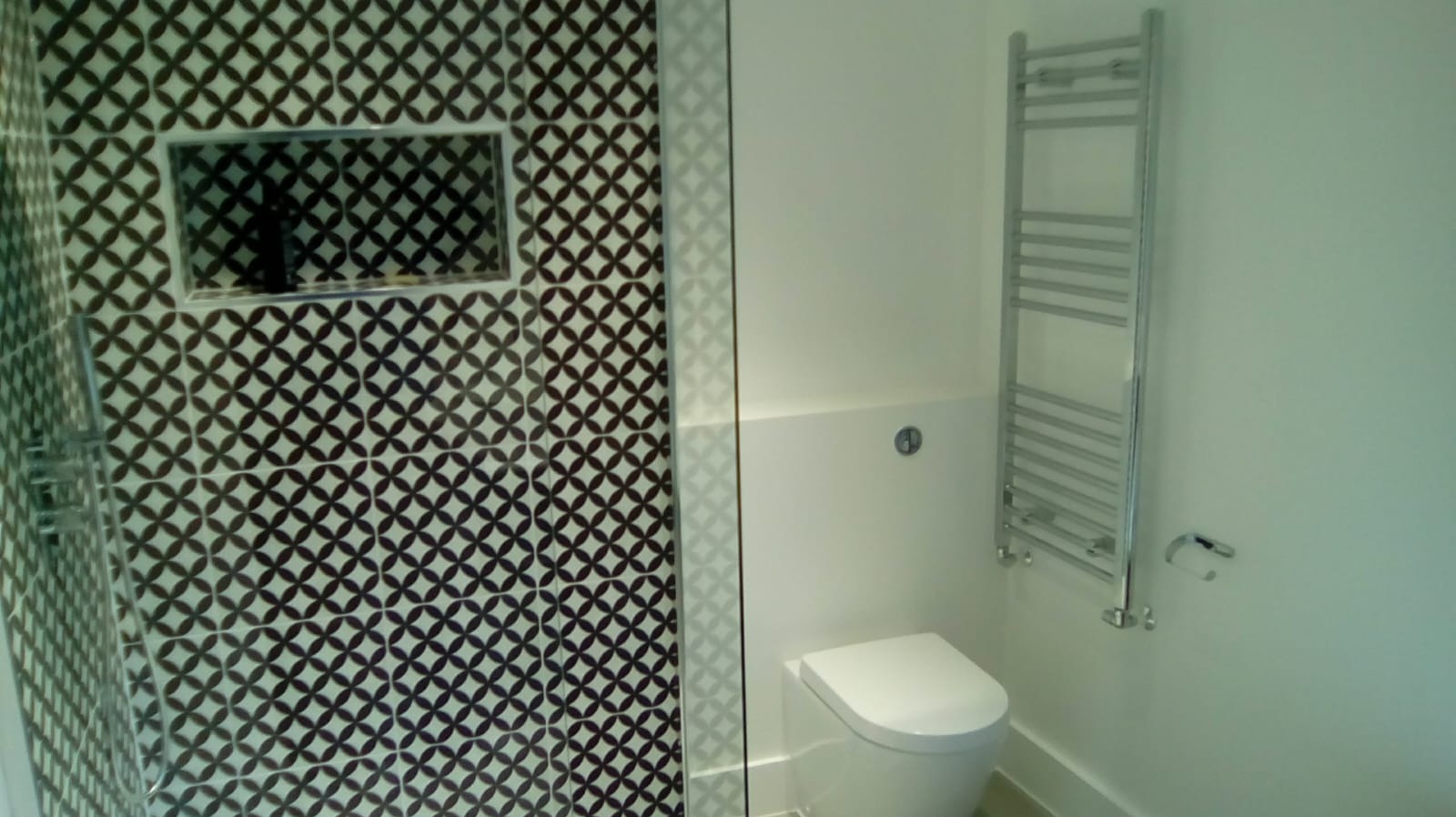 Nowoczesna łazienka z kabiną prysznicową wyłożoną wzorzystymi płytkami w odcieniach brązu i bieli, wiszącą toaletą oraz grzejnikiem drabinkowym.