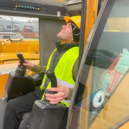Czaju DT - Centrum Szkoleń - Operator w kabinie żółtego dźwigu, ubrany w kask i kamizelkę odblaskową, z uśmiechem spogląda w górę, trzymając dżojstiki sterujące.