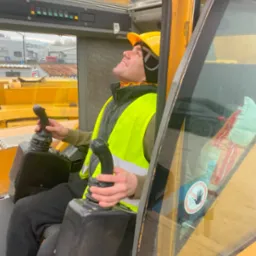 Operator w kabinie żółtego dźwigu, ubrany w kask i kamizelkę odblaskową, z uśmiechem spogląda w górę, trzymając dżojstiki sterujące.