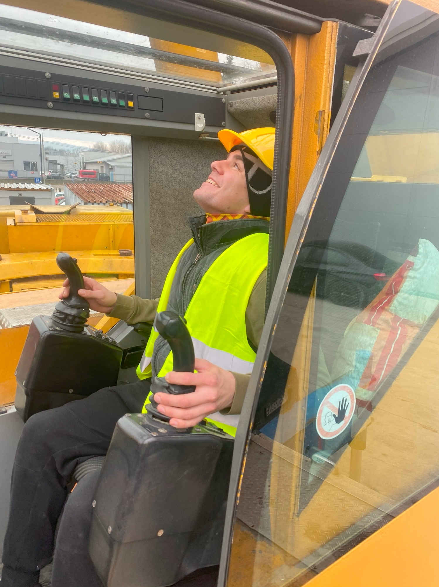 Operator w kabinie żółtego dźwigu, ubrany w kask i kamizelkę odblaskową, z uśmiechem spogląda w górę, trzymając dżojstiki sterujące.
