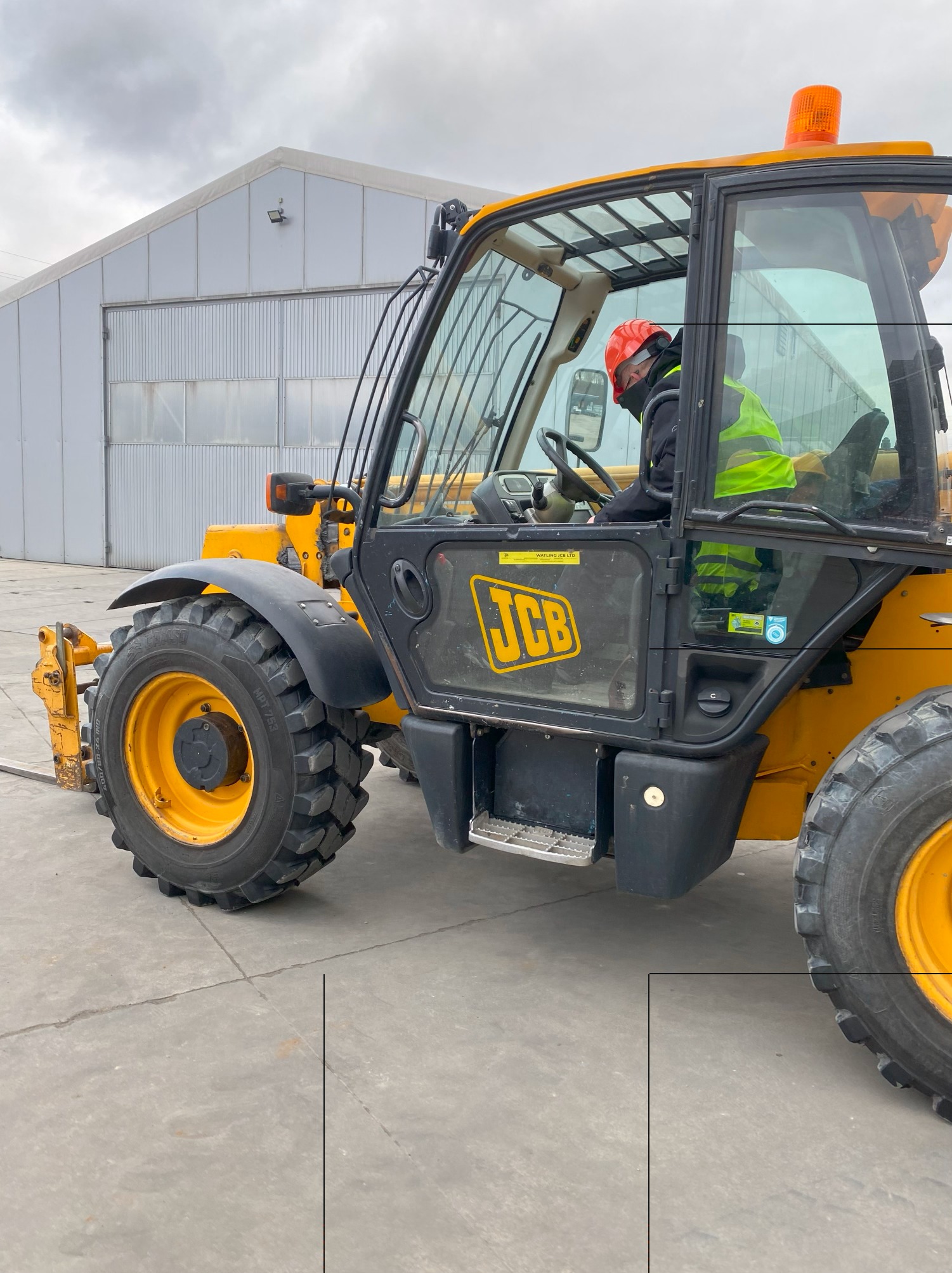 Żółta ładowarka teleskopowa JCB na betonowym placu, operator w kasku i kamizelce odblaskowej w kabinie, widoczny budynek magazynowy w tle.
