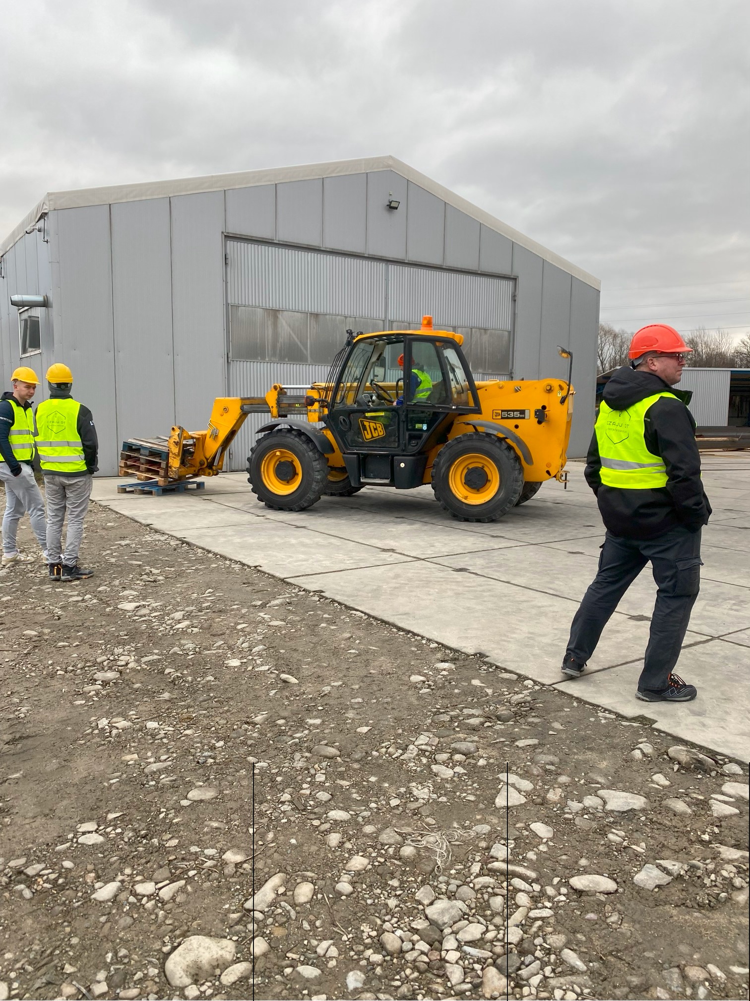 Żółta ładowarka teleskopowa JCB podnosi paletę z pomocą pracowników w odblaskowych kamizelkach na tle szarego magazynu.