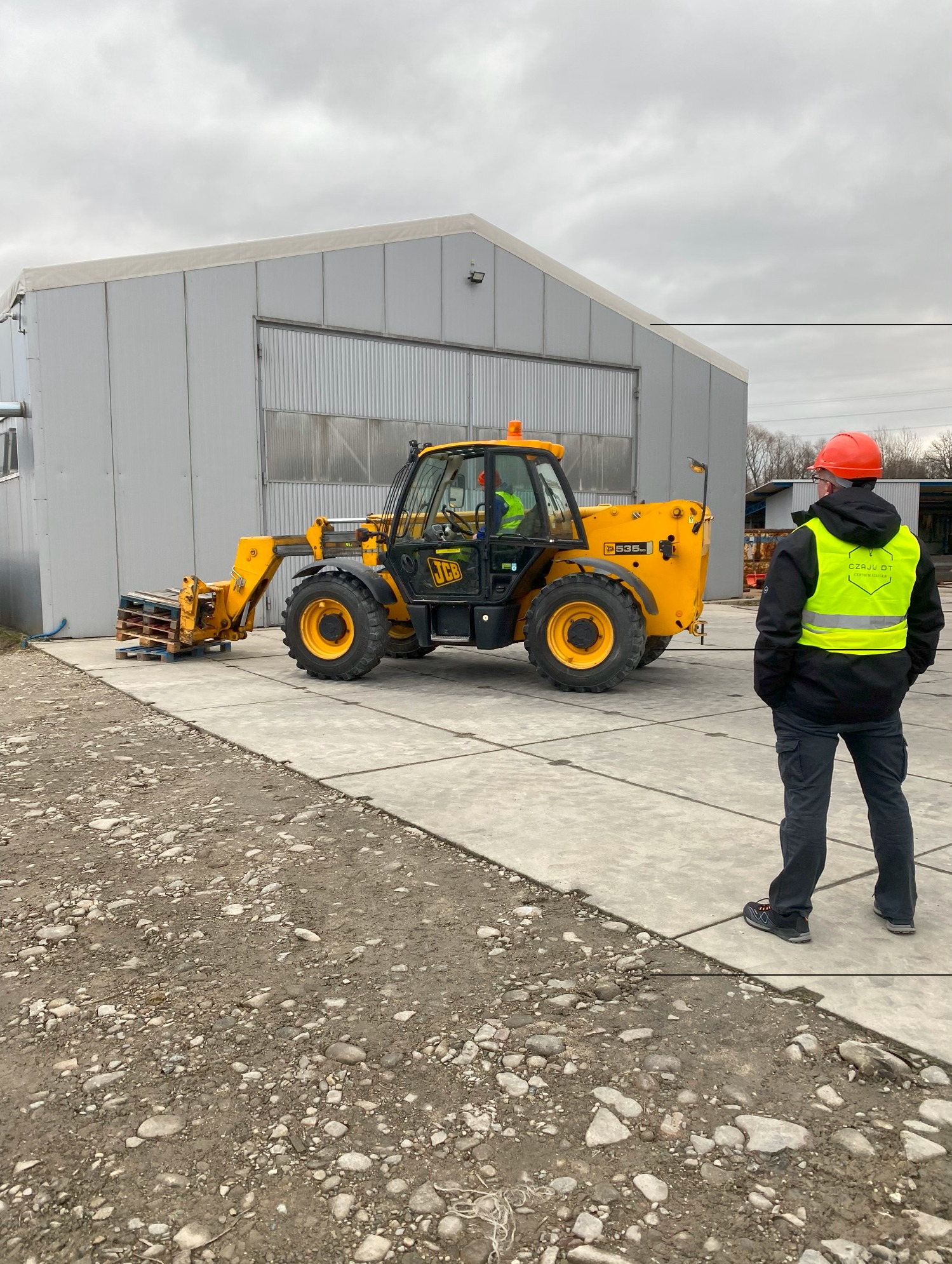 Żółta ładowarka teleskopowa JCB 535-95 z paletami na widłach, operator w kabinie i pracownik w kamizelce odblaskowej i kasku obserwujący pracę na placu budowy.