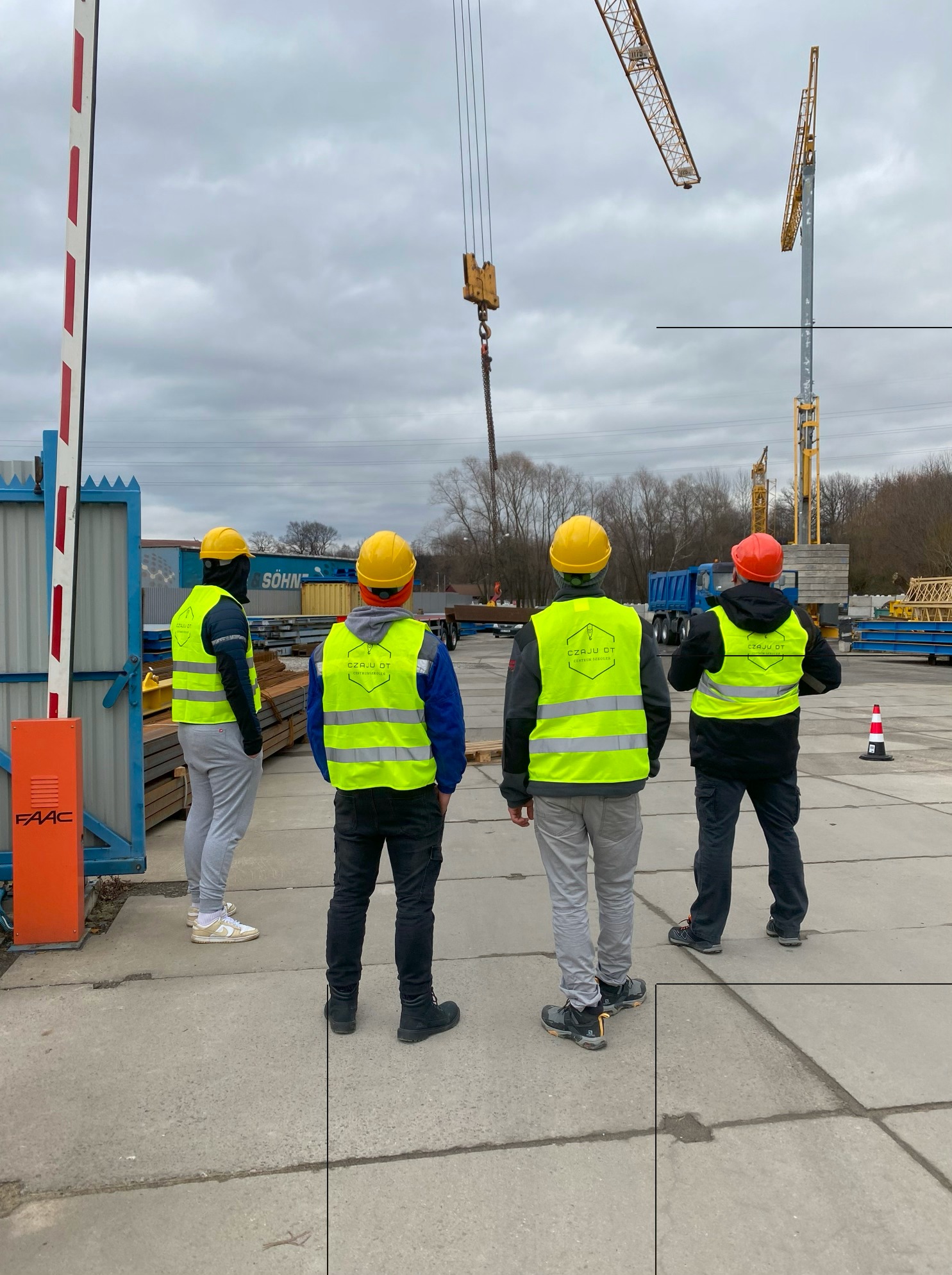 Czterech pracowników w żółtych kamizelkach i kaskach obserwuje pracę dwóch żurawi na placu budowy, z widocznym logo firmy na kamizelkach.