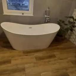 Nowoczesna, wolnostojąca wanna w jasnej łazience z drewnopodobną podłogą i dużym oknem z widokiem na zabudowę jednorodzinną w pochmurny dzień.