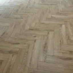 Ułożona podłoga z paneli laminowanych w jodełkę francuską, jasne drewno z widoczną strukturą słojów, delikatne refleksy światła.