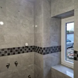 Ściany wyłożone szarą płytką imitującą kamień z dekoracyjnym pasem mozaiki w łazience w Rudnikach, widoczne przyłącza wodne i okno z widokiem na zewnątrz.