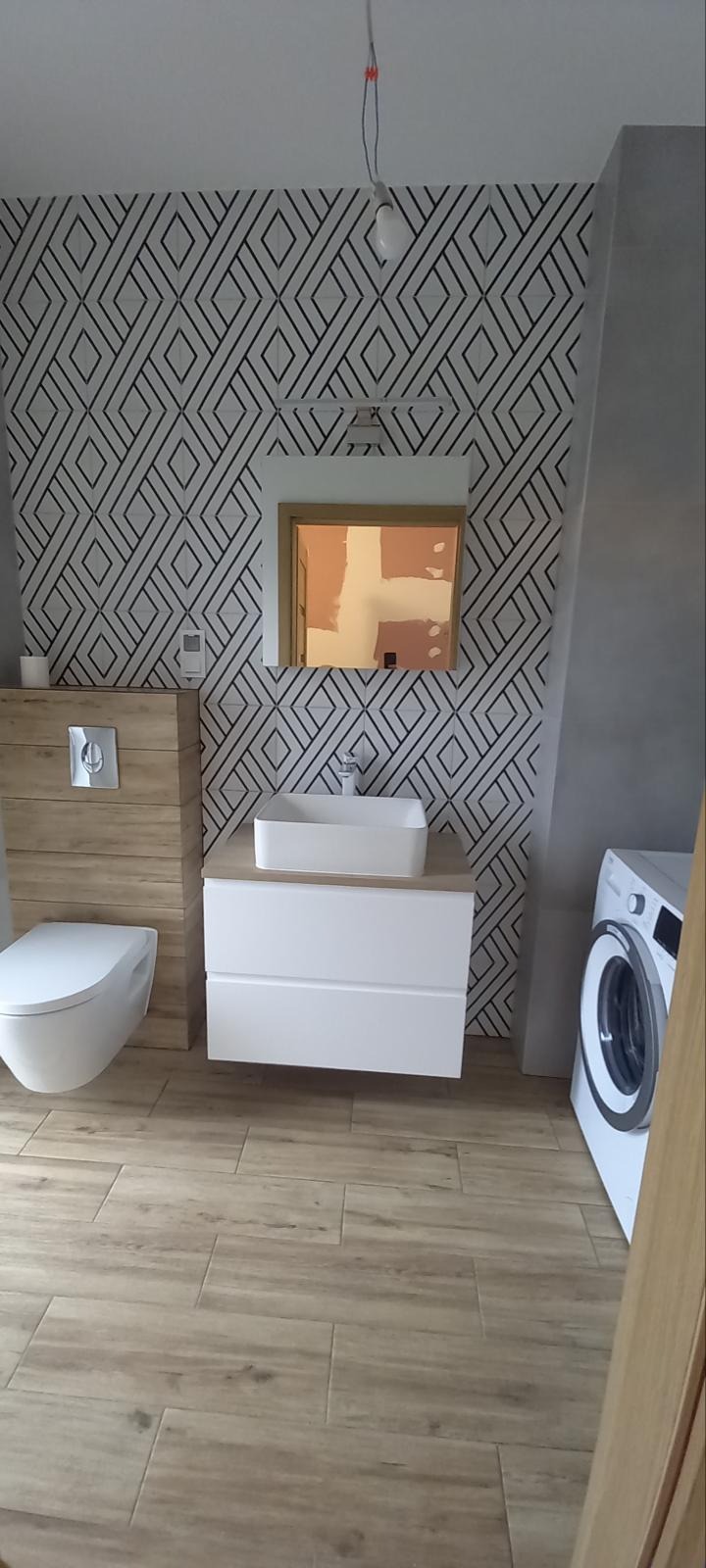 Nowoczesna łazienka z geometryczną tapetą, drewnianą szafką toaletową i podłogą imitującą drewno, z widoczną pralką i niedokończonym fragmentem ściany odbitym w lustrze.