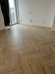 Jasne panele ułożone we wzór jodełki francuskiej w pustym pokoju z białymi ścianami. Widoczne gniazdka elektryczne na ścianie i fragment zasłony.