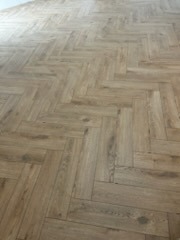 Ułożona podłoga z paneli laminowanych w jodełkę francuską, jasne drewno z widoczną strukturą słojów, delikatne refleksy światła.