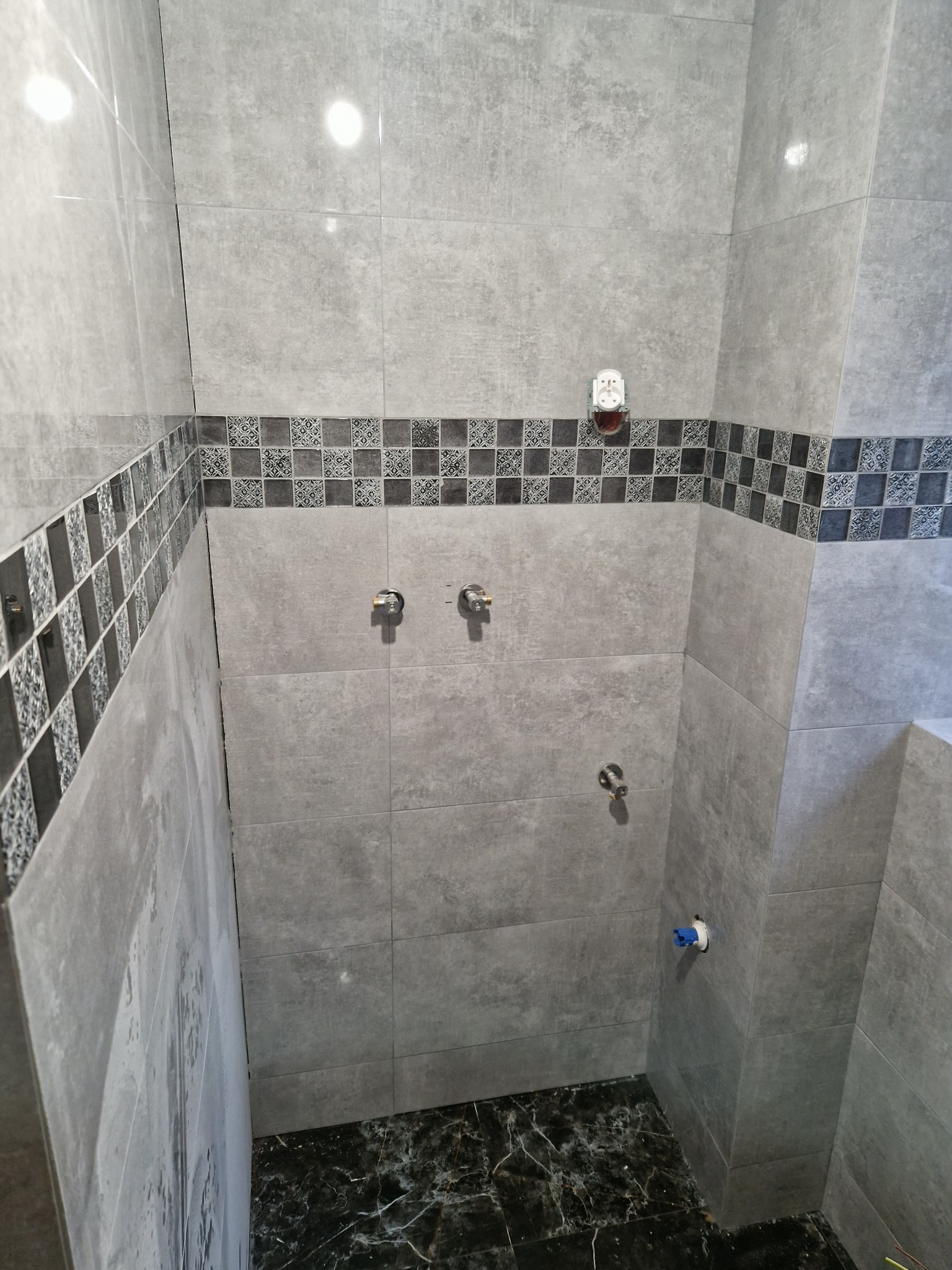 Ściany łazienki wyłożone szarą płytką z dekoracyjnym pasem płytek z wzorem, widoczne przyłącza wodne i gniazdko elektryczne, podłoga z ciemnej płytki imitującej marmur.