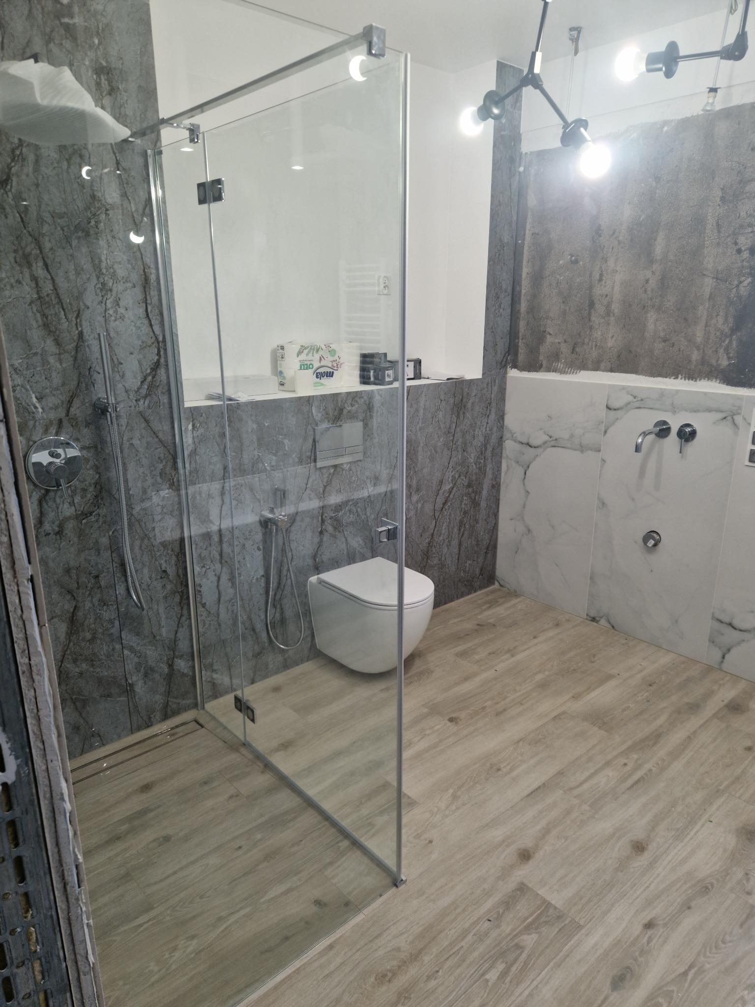 Wnętrze łazienki w trakcie wykańczania z kabiną prysznicową ze szkła, toaletą, marmurowymi płytkami na ścianach i panelami podłogowymi imitującymi drewno, widoczne niedokończone fragmenty ściany.