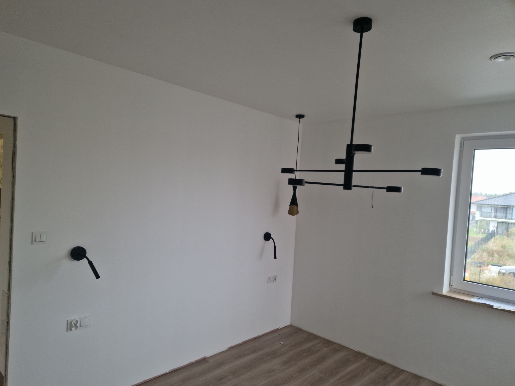 Nowoczesne, czarne oświetlenie sufitowe i ścienne w świeżo wykończonym, jasnym pokoju z panelami podłogowymi i widokiem z okna na zewnątrz.