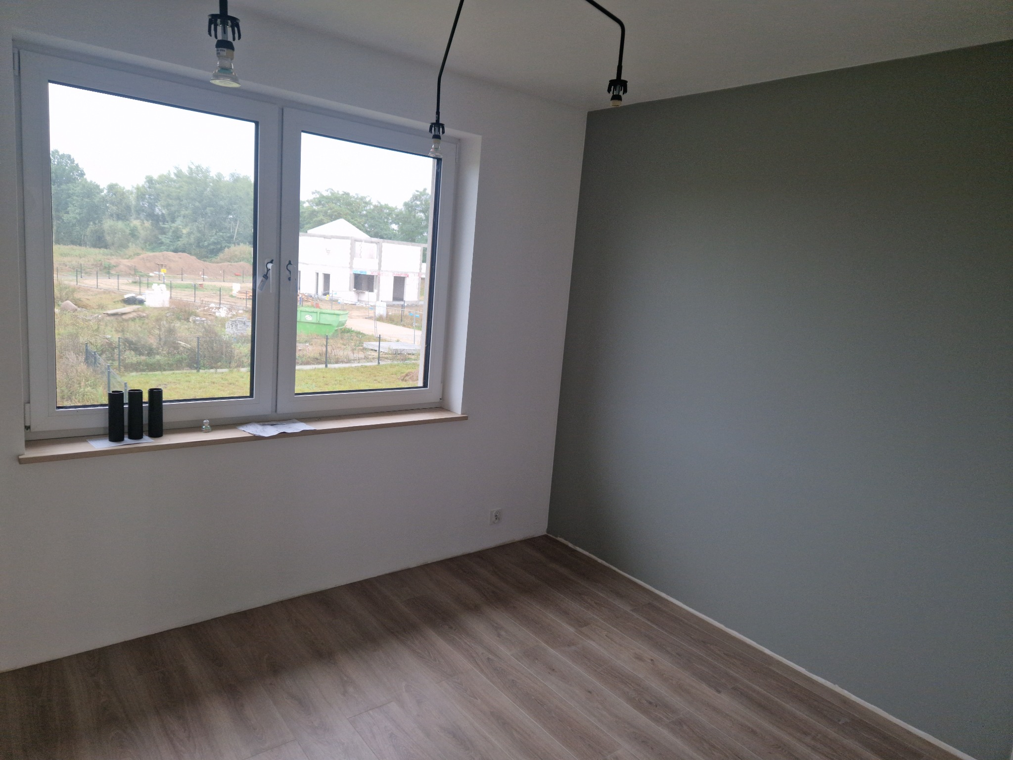 Pusty pokój po remoncie z panelami podłogowymi, szarą ścianą i widokiem na budowę za oknem.