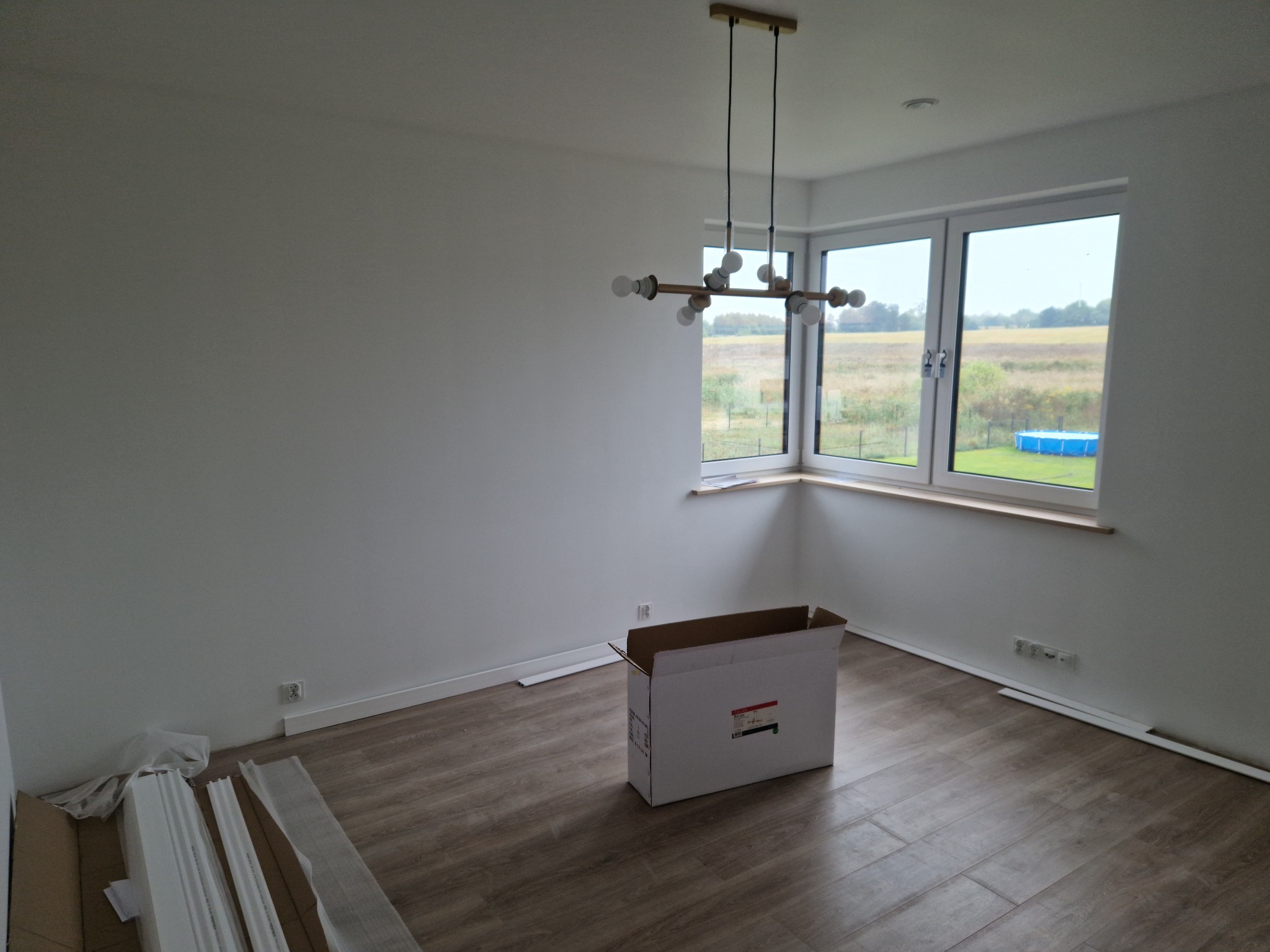 Puste, białe pomieszczenie z drewnopodobną podłogą, z oknami wychodzącymi na krajobraz z basenem, nowoczesna lampa sufitowa, karton i listwy przypodłogowe w opakowaniu leżące na podłodze.