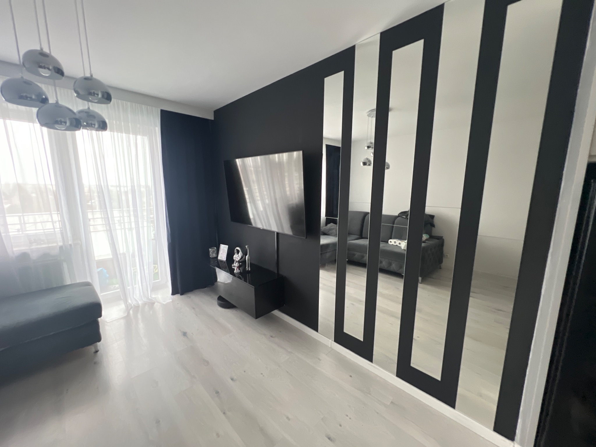 Nowoczesny salon z czarną ścianą, telewizorem, lustrami w czarnych ramach i jasną podłogą, widok na balkon przez firanki.