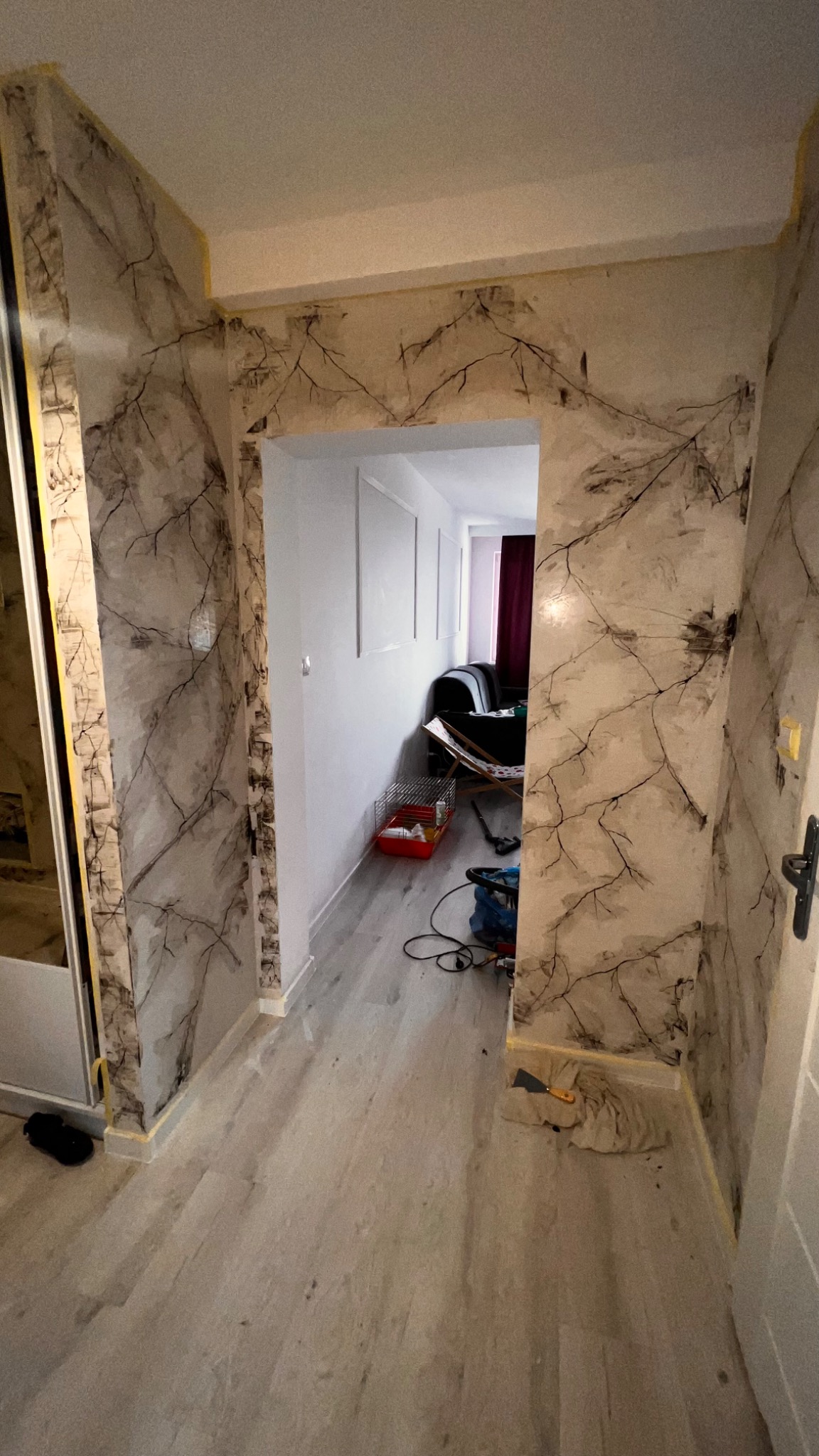 Korytarz po dekoracyjnym malowaniu ścian z motywem gałęzi, zabezpieczone taśmą malarską krawędzie, widoczne narzędzia i ślady pracy na podłodze.