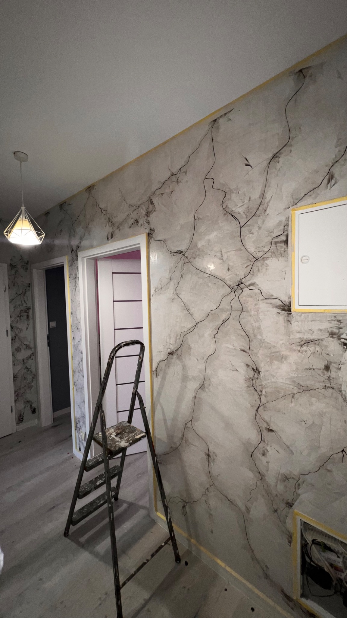 Ściana w pokoju po nałożeniu dekoracyjnej, szarej gładzi imitującej marmur z czarnymi żyłkami, zabezpieczona taśmą malarską, z widoczną, używaną drabiną malarską.